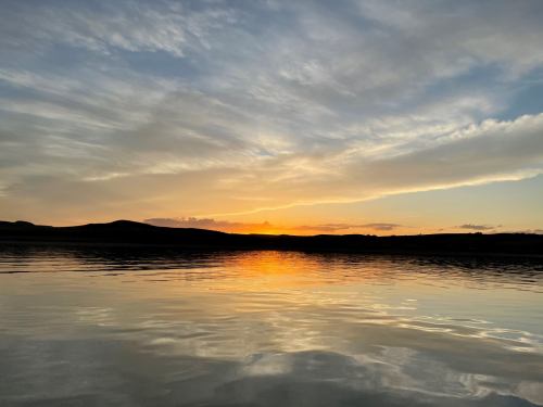 A serene sunset over calm water, with soft clouds and silhouettes of hills in the background.