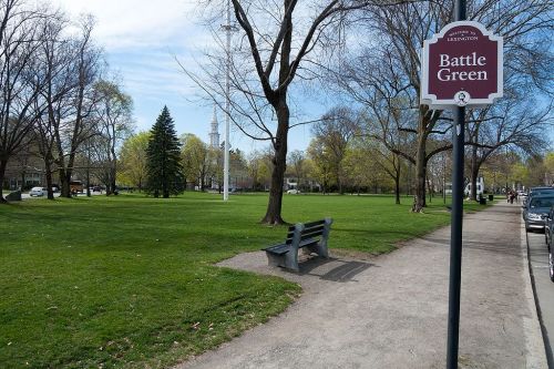 A park scene featuring a sign for Battle Green, with trees, grass, and a bench along a pathway.