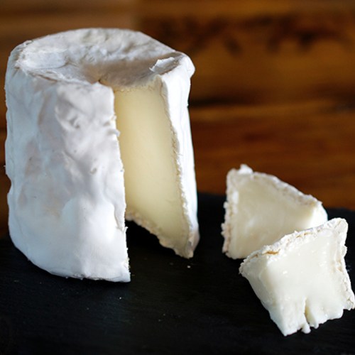 A round wheel of soft cheese with a white rind, partially cut, on a dark slate surface.