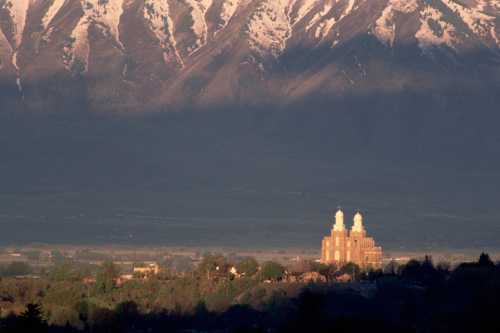 A majestic mountain range with snow-capped peaks towers over a valley, featuring a distant temple surrounded by greenery.