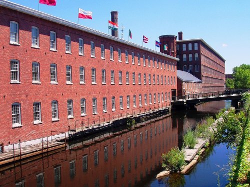 Historic red brick mill building alongside a calm canal, with flags flying and reflections in the water.