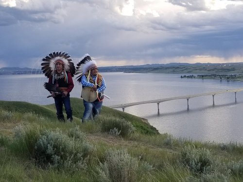 Two individuals in traditional Native American attire walk along a grassy hill by a lake, with a bridge in the background.