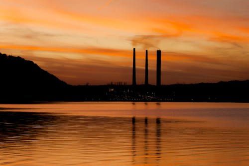 Silhouetted power plant towers against a colorful sunset, reflecting on calm water.