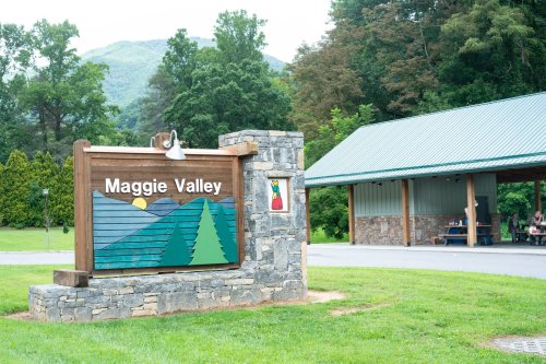 Sign for Maggie Valley with mountains and trees, next to a covered area and lush greenery in the background.