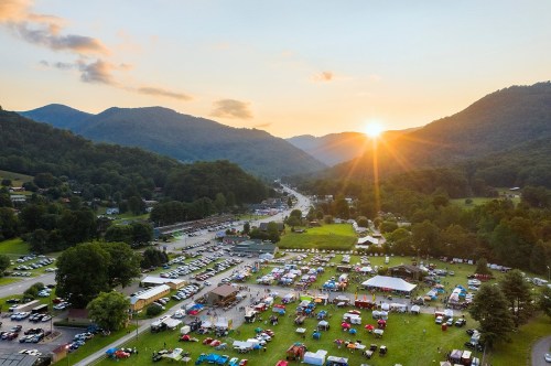 Aerial view of a festival at sunset, surrounded by mountains, with tents and cars in a grassy area.