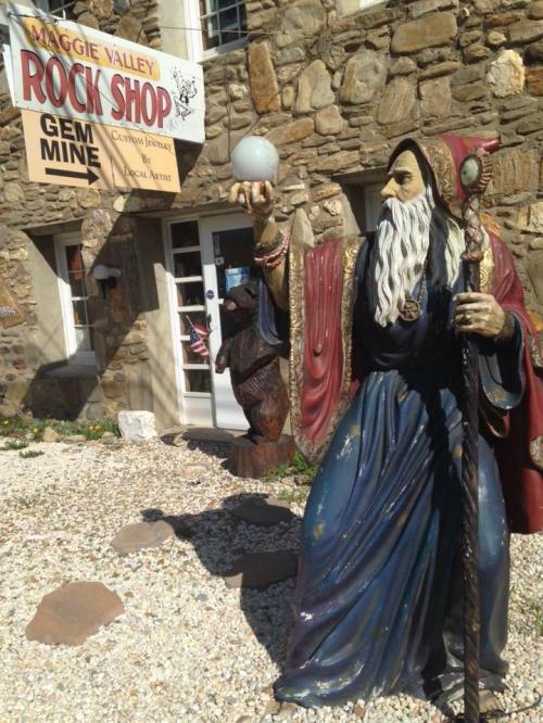 A wizard statue holding a crystal ball stands outside the Maggie Valley Rock Shop, featuring a stone building and sign.