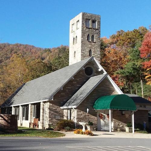 A stone church with a green awning, surrounded by colorful autumn trees and a tall tower in the background.