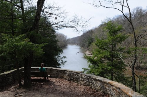 A person sits on a stone bench overlooking a serene river surrounded by trees in a peaceful natural setting.