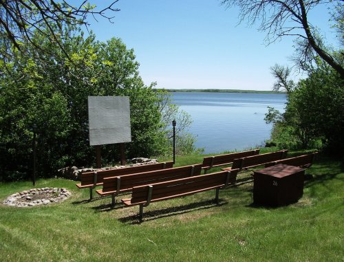 A serene lakeside scene with benches facing a calm water view, surrounded by greenery and a fire pit nearby.