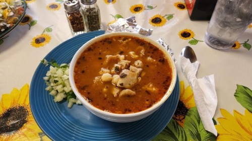 A bowl of hearty soup with chicken, spices, and toppings, served on a blue plate with diced onions on the side.
