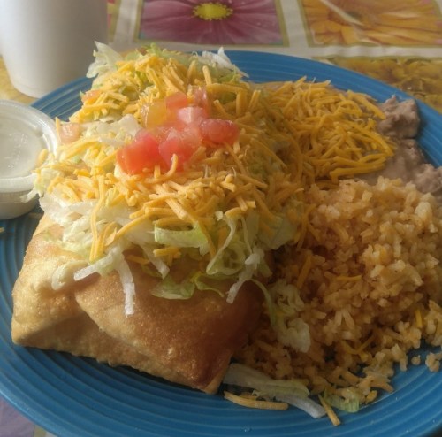 A blue plate with a crispy taco topped with lettuce, cheese, and tomatoes, served with rice and refried beans.