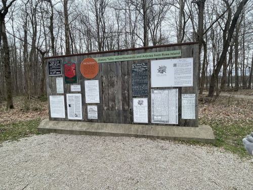A wooden information board in a forest, displaying various historical articles and documents about Rose Island.