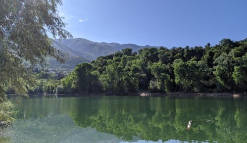 A serene lake surrounded by lush greenery and mountains under a clear blue sky. Reflections shimmer on the water's surface.