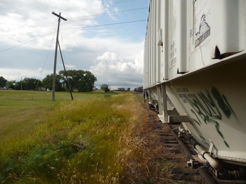 A view along a train track with a freight car on the left, grassy fields, and a cloudy sky in the background.