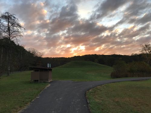 A winding path leads to a grassy hill under a colorful sunset with clouds in the sky.