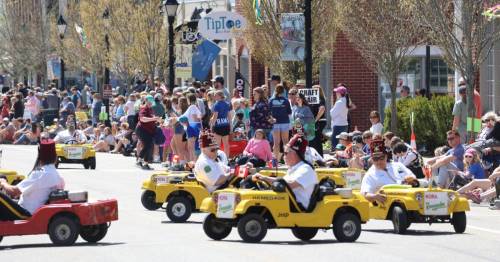 A parade scene with people watching as small cars drive by, featuring participants in costumes and festive decorations.