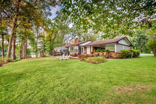 A cozy house surrounded by trees and a lush green lawn, featuring a patio with chairs and a peaceful outdoor setting.