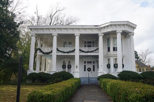 A large white historic house with columns, decorated with greenery, surrounded by manicured bushes and a cloudy sky.