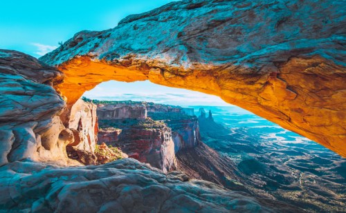 A stunning view through a natural rock arch, overlooking colorful cliffs and a vast landscape under a bright blue sky.