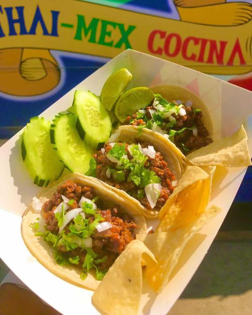 Three tacos filled with meat, topped with onions and cilantro, served with cucumber slices, lime wedges, and tortilla chips.