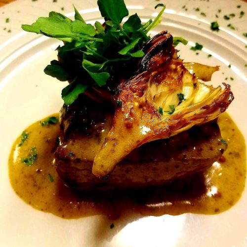 A beautifully plated dish featuring a piece of meat topped with greens and a roasted artichoke, drizzled with sauce.