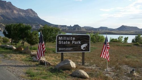Sign for Millsite State Park with American flags, surrounded by rocky hills and a lake in the background.