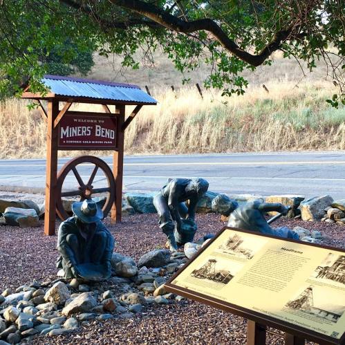 Welcome sign for Mines' Bend with bronze sculptures of miners and an informational plaque in a natural setting.
