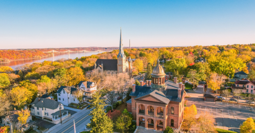 Aerial view of a scenic town with colorful autumn trees, a church spire, and a river in the background.