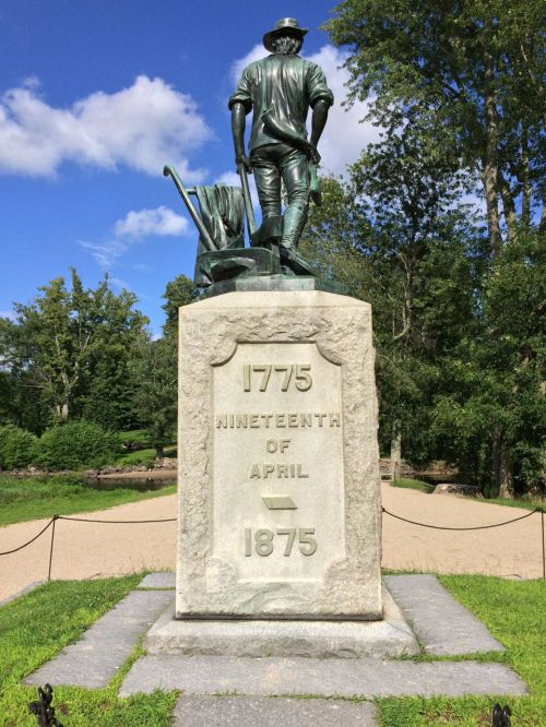 Statue of a farmer with a plow, commemorating April 19, 1775, and 1875, surrounded by trees and blue sky.