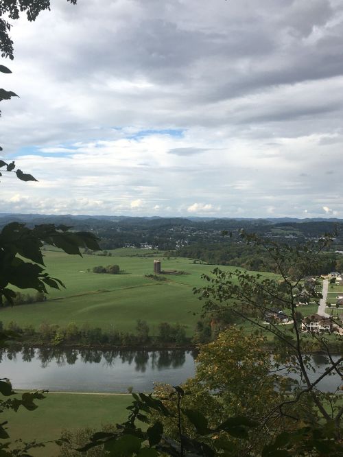 A scenic view of green fields, a river, and distant mountains under a cloudy sky, framed by tree branches.