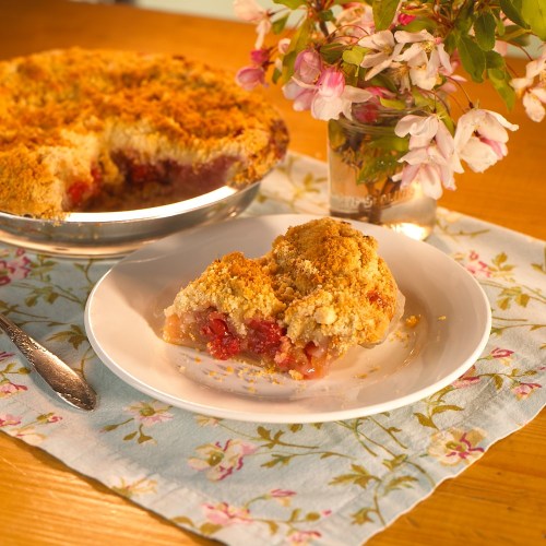 A slice of cherry pie on a plate beside a vase of flowers, with the pie in a dish on a floral tablecloth.