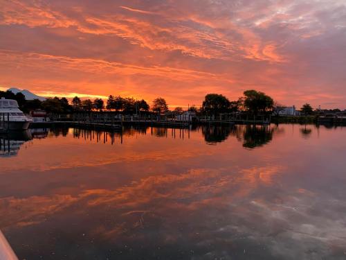 A vibrant sunset reflects on calm water, with silhouettes of trees and boats along the shore.