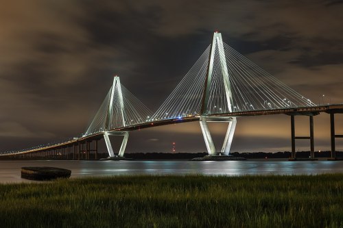 A illuminated cable-stayed bridge at night, reflecting in calm waters with grassy banks in the foreground.