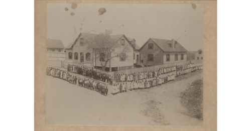 Historic black-and-white photo of a large group of people gathered outside a building, with trees and houses in the background.