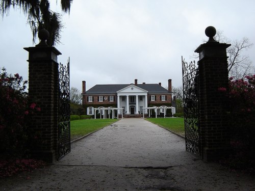 A grand brick mansion with white columns, viewed through ornate gates, surrounded by lush greenery and blooming flowers.