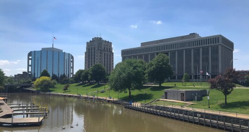 Cityscape featuring modern buildings along a river, with green lawns and trees under a clear blue sky.