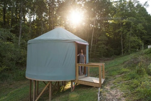A woman stands at the entrance of a yurt surrounded by trees, with the sun setting in the background.