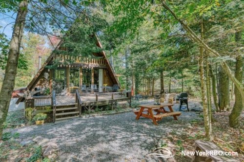 A cozy A-frame cabin surrounded by trees, featuring a deck, picnic table, and outdoor seating area.