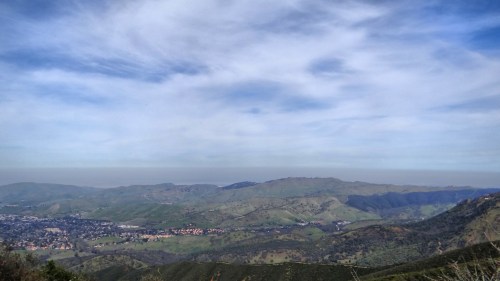 A panoramic view of rolling green hills under a cloudy sky, with a distant city visible in the valley below.