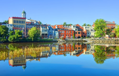 Colorful buildings reflect in calm water under a clear blue sky, creating a picturesque riverside scene.