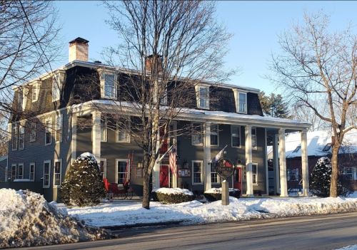A large, historic house with a porch, surrounded by snow and trees, featuring an American flag and a welcoming entrance.