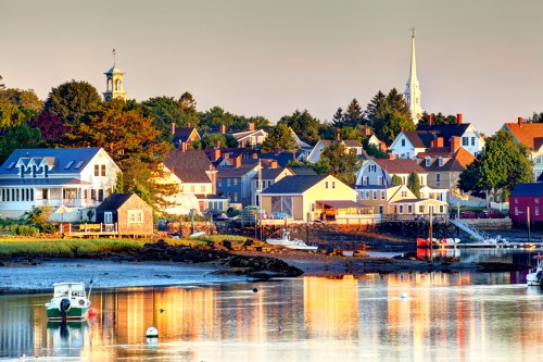 A picturesque coastal town at sunrise, featuring colorful houses, a church steeple, and calm waters reflecting the scenery.