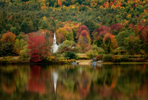 A serene lake reflects vibrant autumn foliage and a white church steeple amidst colorful trees.