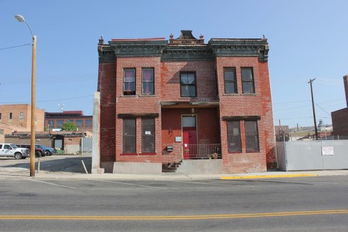 A two-story brick building with red windows and a decorative roof, located on a street with parked cars nearby.