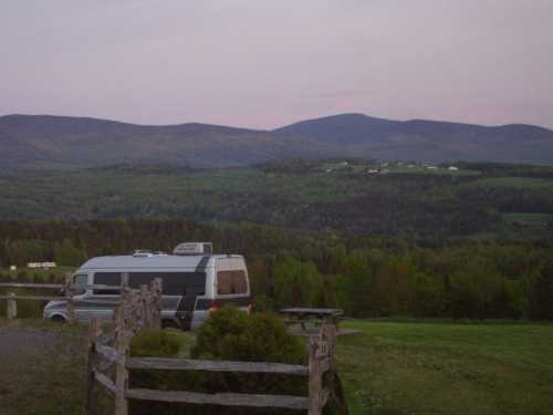 A camper van parked near a wooden fence, with rolling green hills and mountains in the background at dusk.