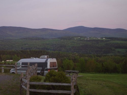 A camper van parked near a wooden fence, with rolling green hills and mountains in the background at dusk.