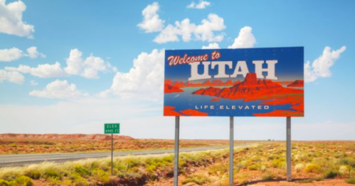 A colorful "Welcome to Utah" sign against a blue sky with clouds, featuring red rock formations in the background.