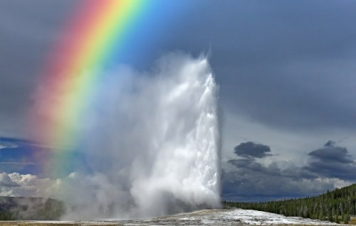 A vibrant rainbow arcs over a geyser erupting in a scenic landscape with clouds and trees in the background.