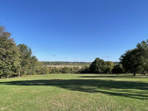 A wide, open grassy field under a clear blue sky, surrounded by trees and distant hills.