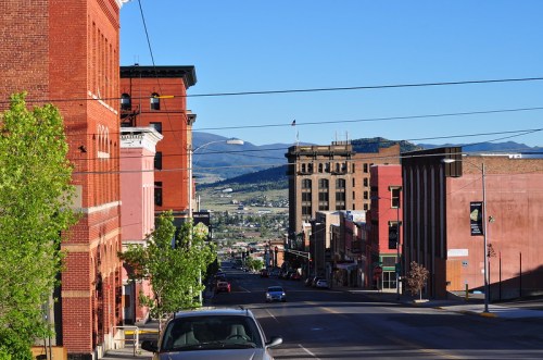 A scenic view of a street lined with historic buildings, leading down to a valley and mountains in the background.
